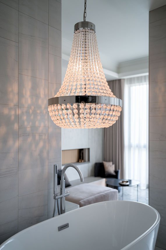 Modern crystal chandelier above bathtub with gray tiles and minimalist decor.